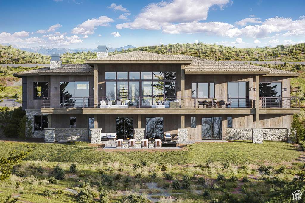 Back of property with stone siding, a balcony, a chimney, a patio, and a lawn