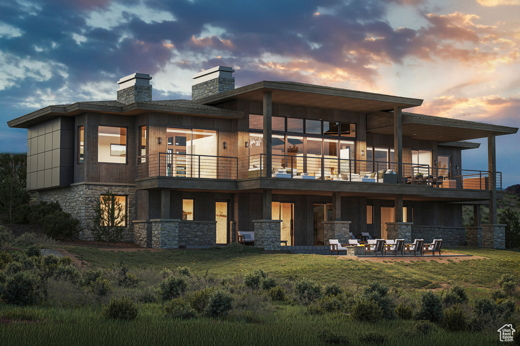 Back of property at dusk with a balcony, stone siding, a patio area, a yard, and a chimney