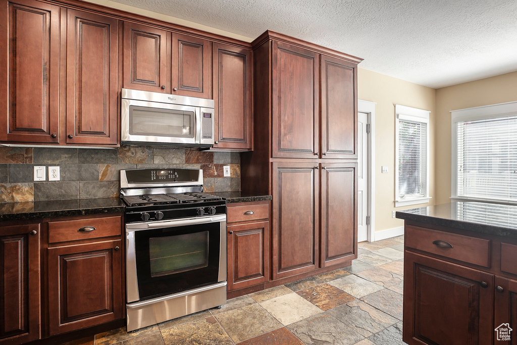 Kitchen featuring appliances with stainless steel finishes, dark countertops, decorative backsplash, stone tile flooring, and baseboards