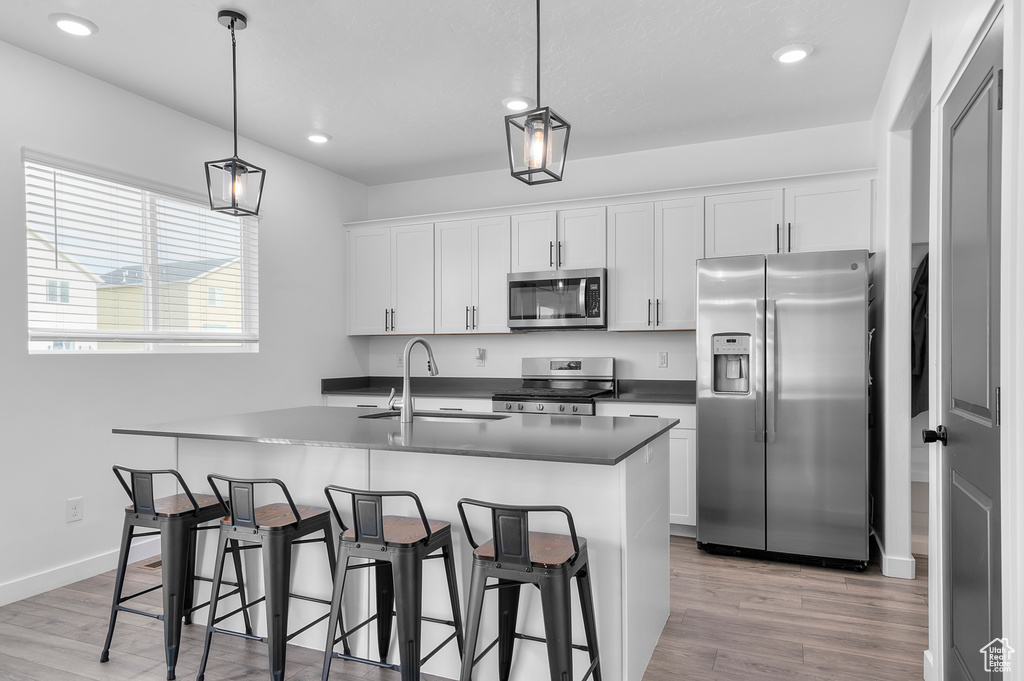 Kitchen featuring stainless steel appliances, a sink, light wood finished floors, dark countertops, and an island with sink