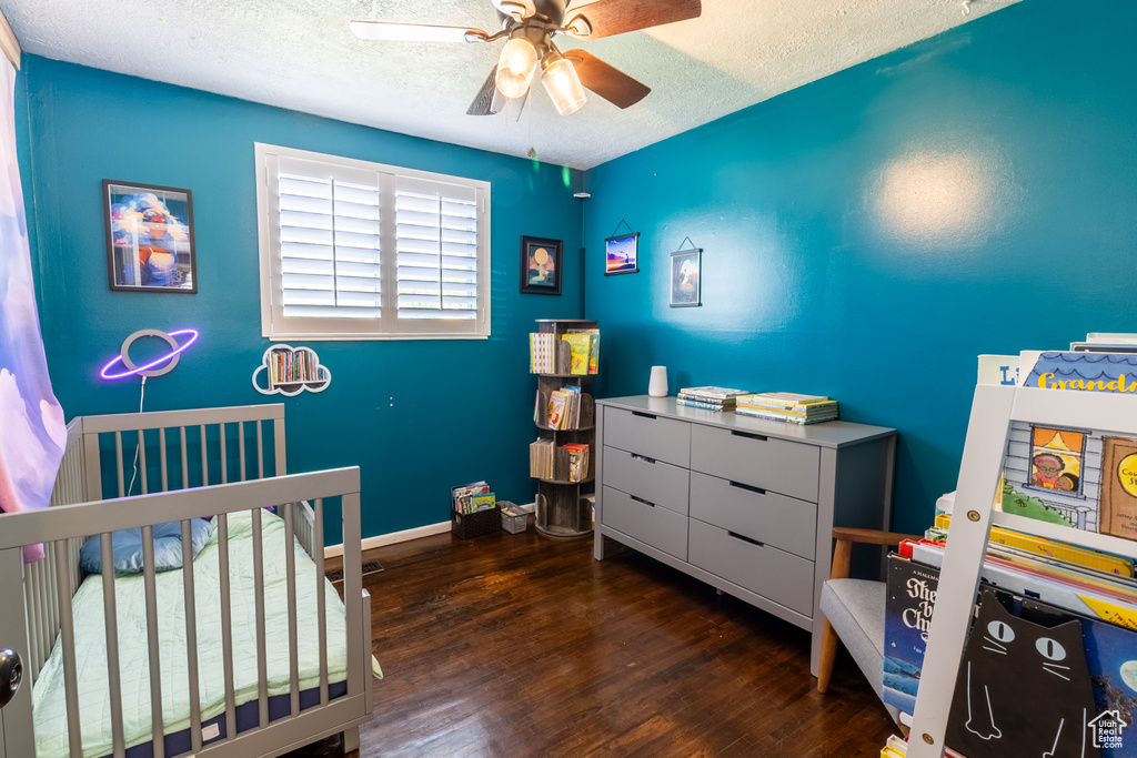 Bedroom with a textured ceiling, wood finished floors, a crib, ceiling fan, and baseboards