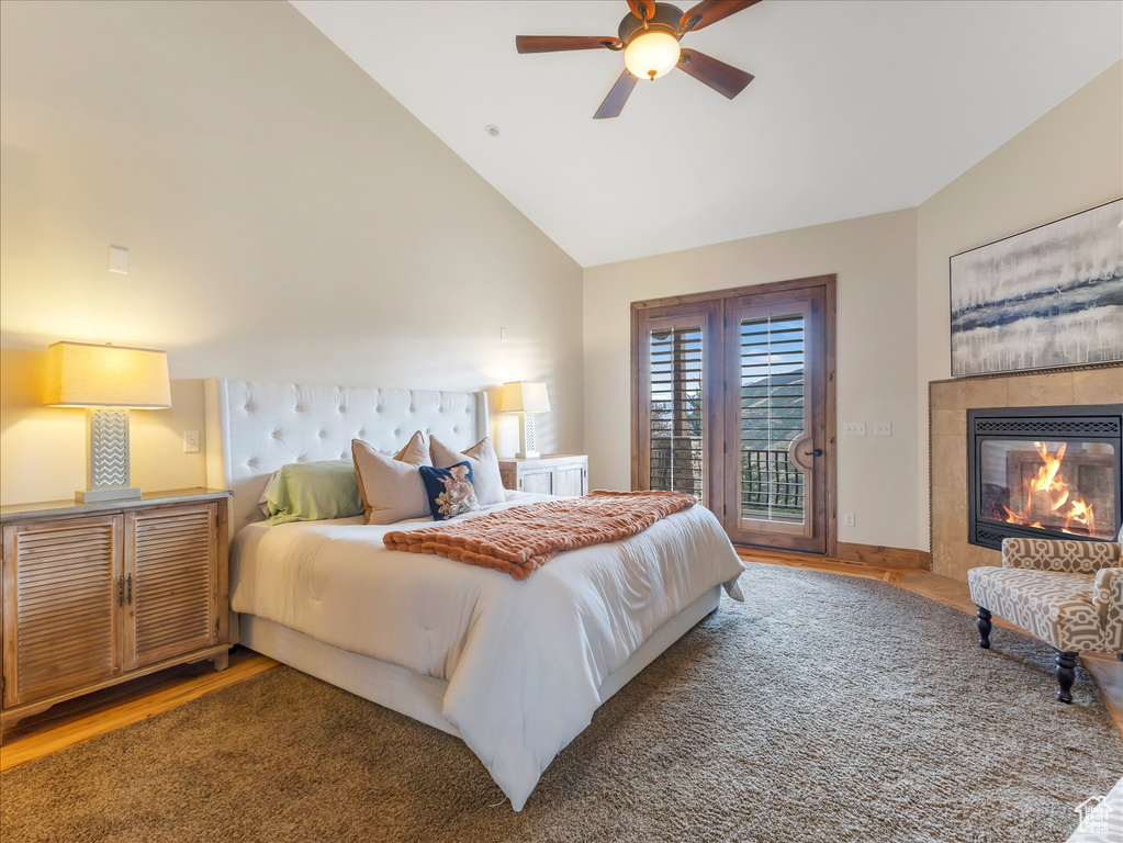 Bedroom with access to exterior, wood finished floors, a tiled fireplace, ceiling fan, and high vaulted ceiling