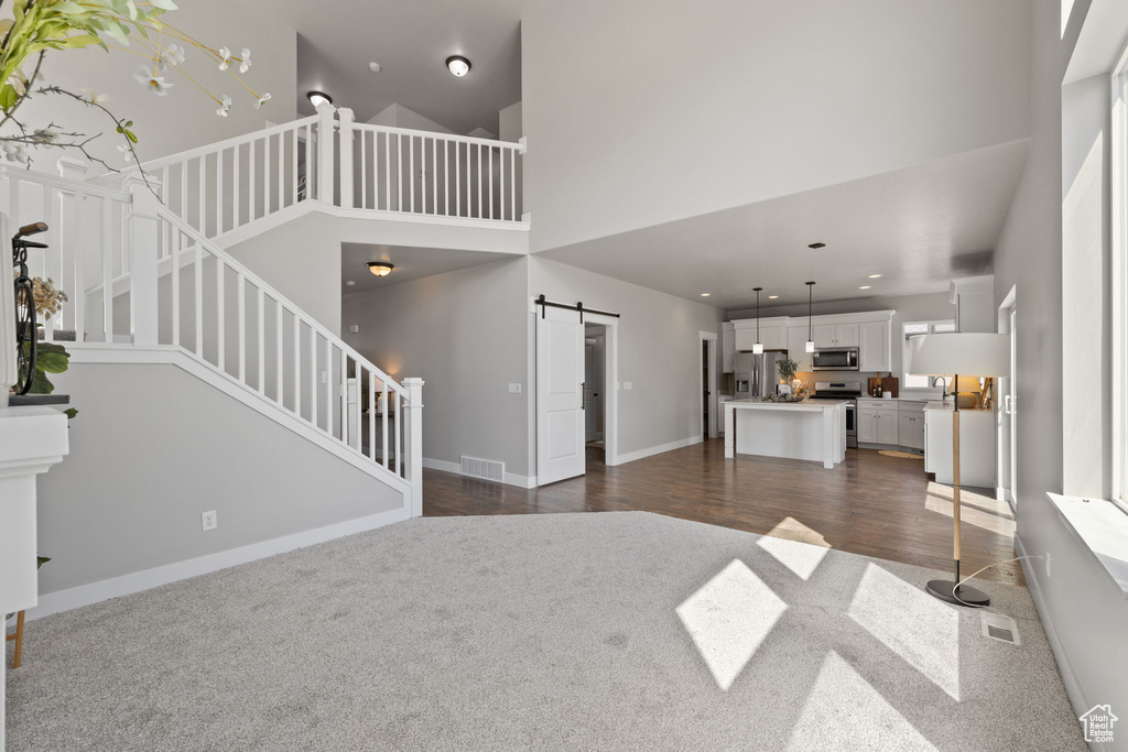 Unfurnished living room with a barn door, dark colored carpet, a towering ceiling, stairs, and baseboards