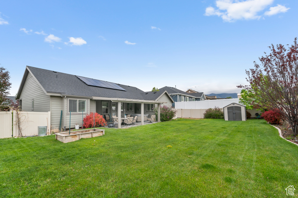 Rear view of property with a storage unit, a patio, roof mounted solar panels, an outbuilding, and a shingled roof