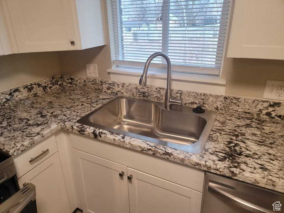 Kitchen view of dishwasher, white cabinets, a sink, and light stone counters