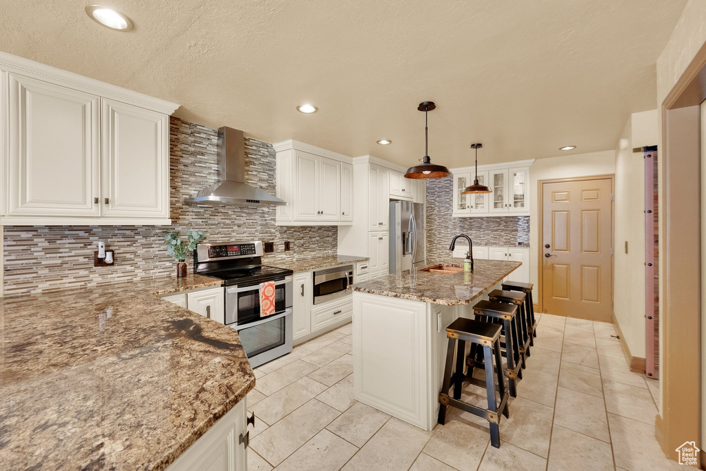 Kitchen with wall chimney range hood, appliances with stainless steel finishes, a sink, decorative backsplash, and light stone counters