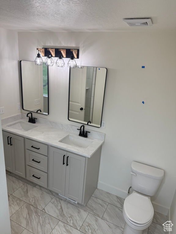 Bathroom featuring a textured ceiling, toilet, marble look tile flooring, double vanity, and baseboards