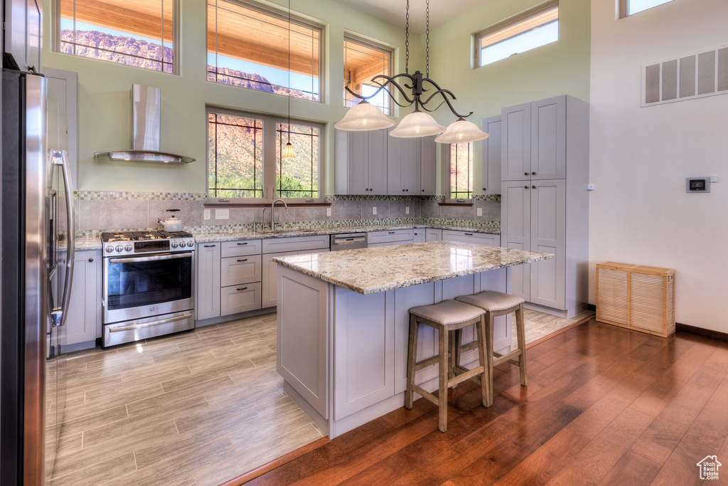 Kitchen featuring stainless steel appliances, wall chimney range hood, a towering ceiling, a sink, and light wood-type flooring