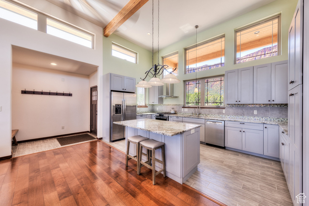 Kitchen with appliances with stainless steel finishes, a towering ceiling, wall chimney range hood, light wood-style floors, and a sink