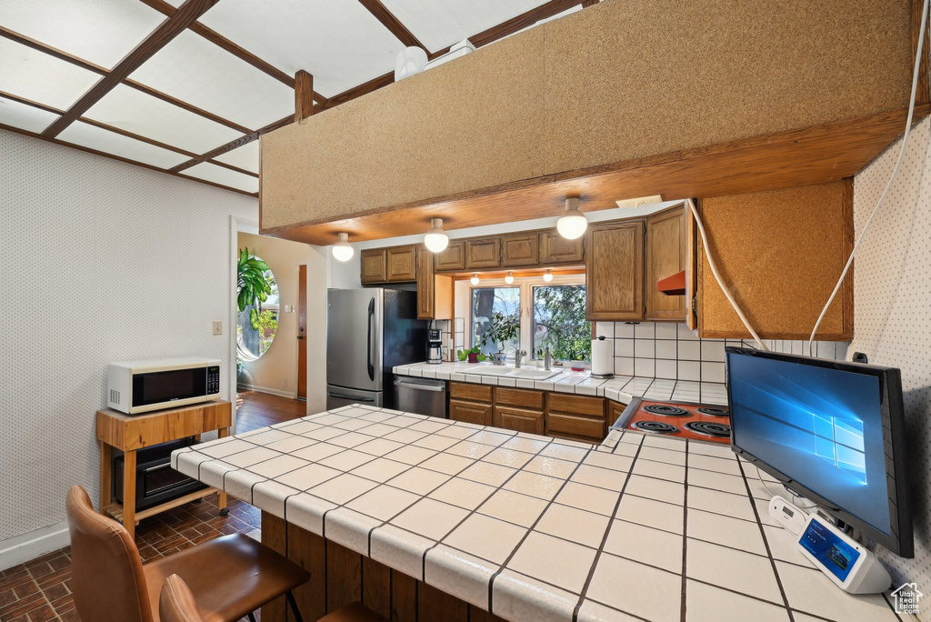 Kitchen featuring stainless steel appliances, tile counters, baseboards, brown cabinetry, and wallpapered walls