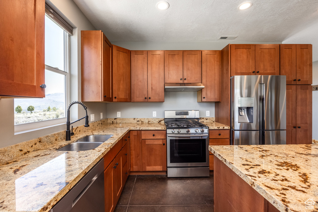 Kitchen featuring stainless steel appliances, a sink, under cabinet range hood, light stone counters, and brown cabinetry