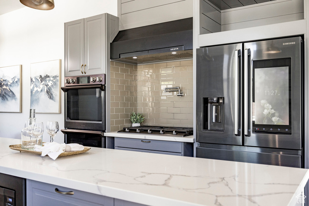 Kitchen featuring stainless steel appliances, gray cabinets, wall chimney exhaust hood, backsplash, and light stone counters