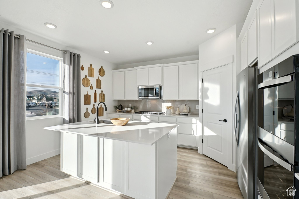 Kitchen featuring appliances with stainless steel finishes, a center island with sink, tasteful backsplash, light countertops, and white cabinets