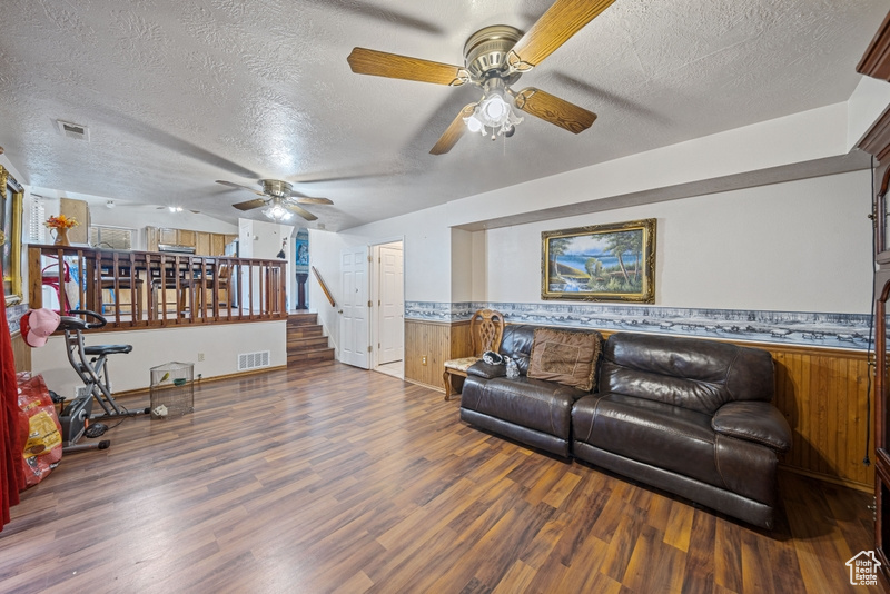 Living area featuring ceiling fan, a textured ceiling, wood finished floors, wainscoting, and wooden walls