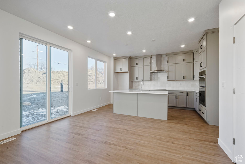 Kitchen featuring wall chimney range hood, gray cabinets, double oven, a kitchen island with sink, and baseboards