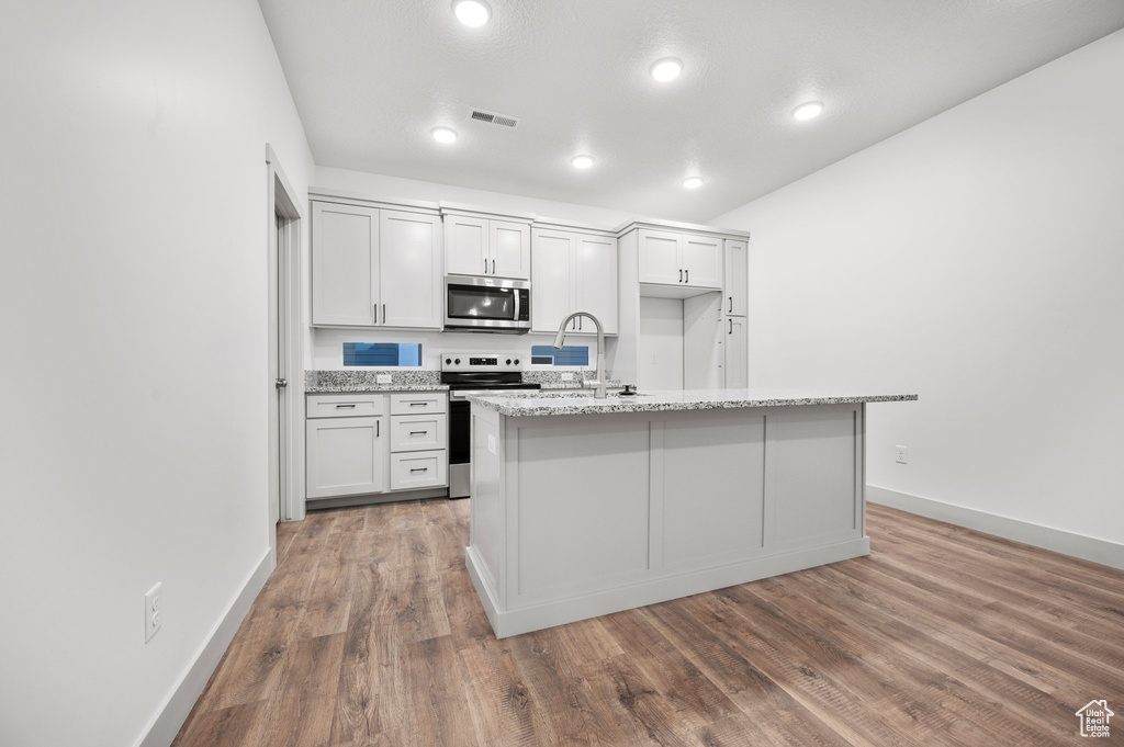 Kitchen with stainless steel appliances, wood finished floors, a kitchen island with sink, baseboards, and light stone countertops