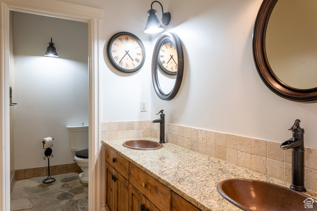 Full bath featuring toilet, double vanity, decorative backsplash, stone finish floors, and baseboards