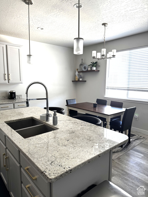 Kitchen with a sink, an island with sink, wood finished floors, a textured ceiling, and hanging light fixtures