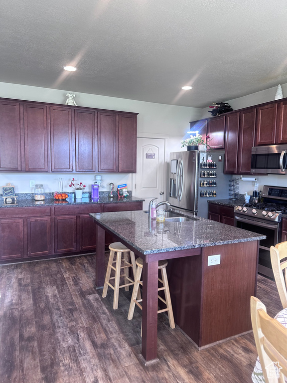 Kitchen featuring appliances with stainless steel finishes, a sink, dark wood-type flooring, dark stone countertops, and a breakfast bar