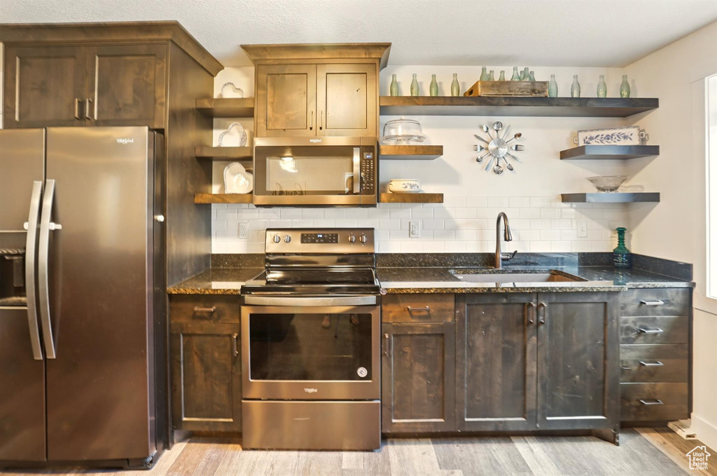 Kitchen with stainless steel appliances, open shelves, a sink, light wood-style floors, and dark stone counters