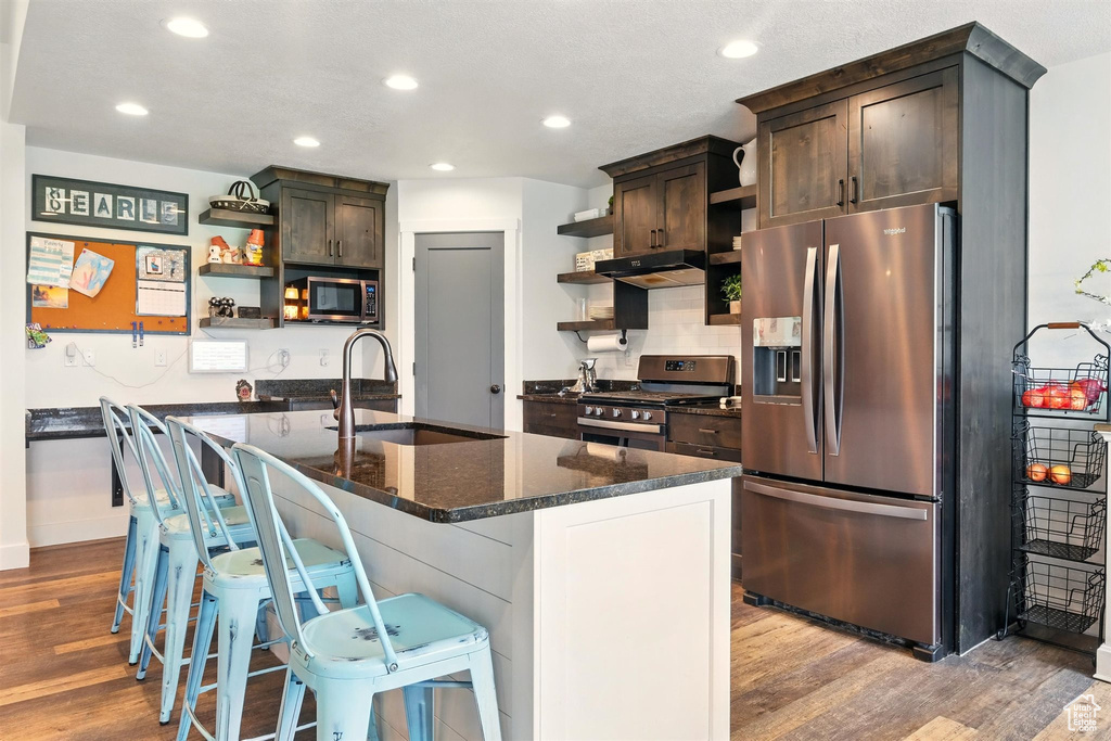 Kitchen with open shelves, stainless steel appliances, under cabinet range hood, wood finished floors, and dark brown cabinetry
