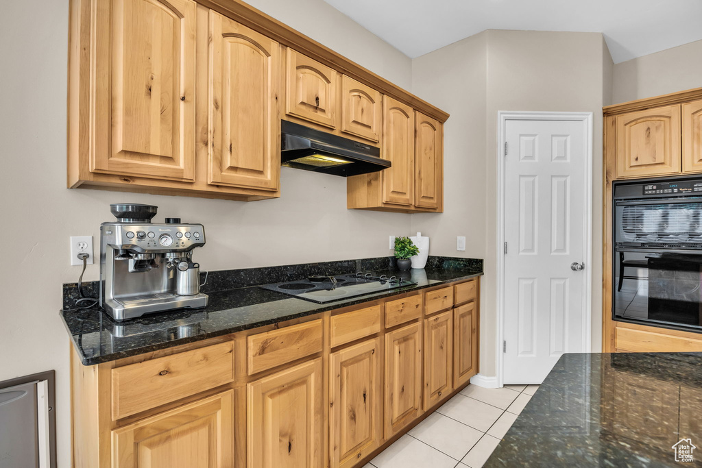 Kitchen featuring under cabinet range hood, white cooktop, light tile patterned flooring, and dark stone counters