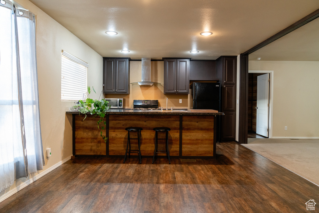 Kitchen with wall chimney exhaust hood, black appliances, a peninsula, dark wood-style flooring, and recessed lighting