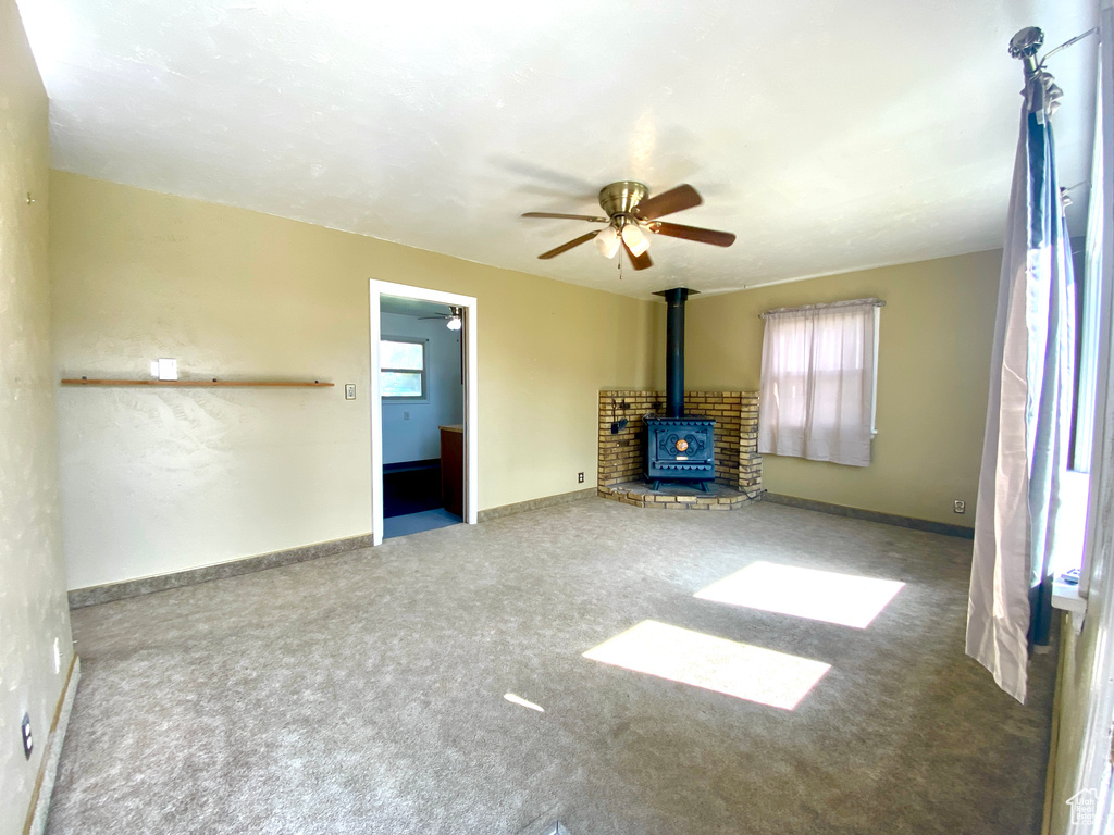 Unfurnished living room featuring a wood stove, carpet flooring, a ceiling fan, and baseboards