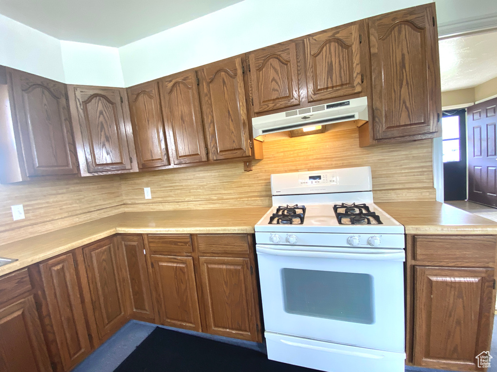 Kitchen with white gas stove, under cabinet range hood, light countertops, and backsplash
