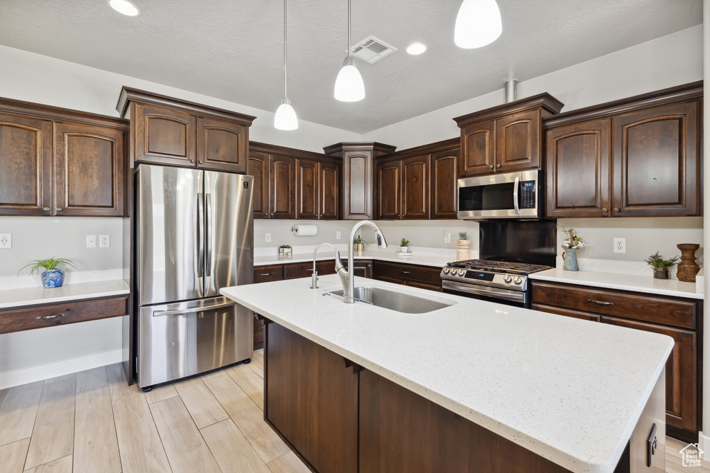 Kitchen featuring stainless steel appliances, a sink, pendant lighting, dark brown cabinets, and an island with sink