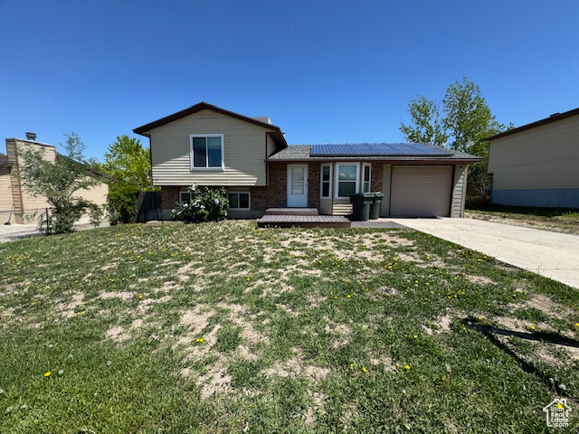 Split level home with brick siding, driveway, roof mounted solar panels, an attached garage, and a front lawn