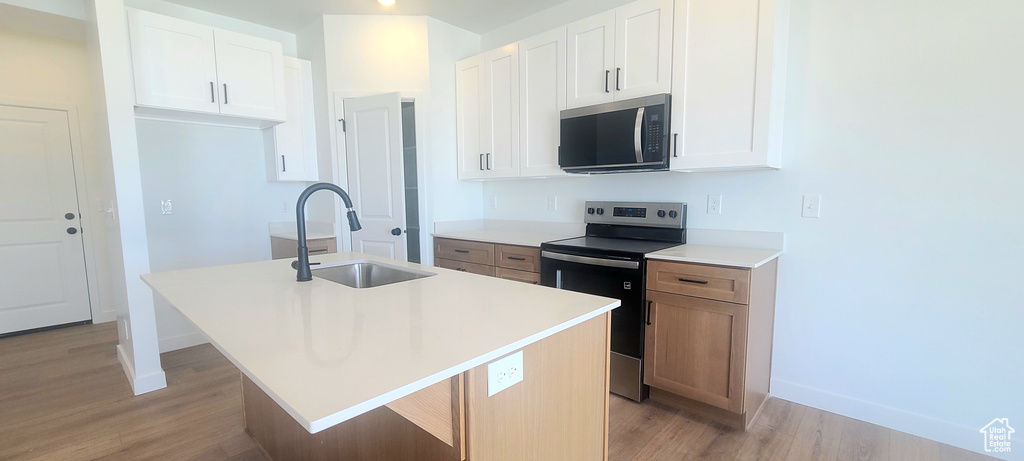 Kitchen with stainless steel appliances, a sink, light wood-style flooring, a center island with sink, and baseboards