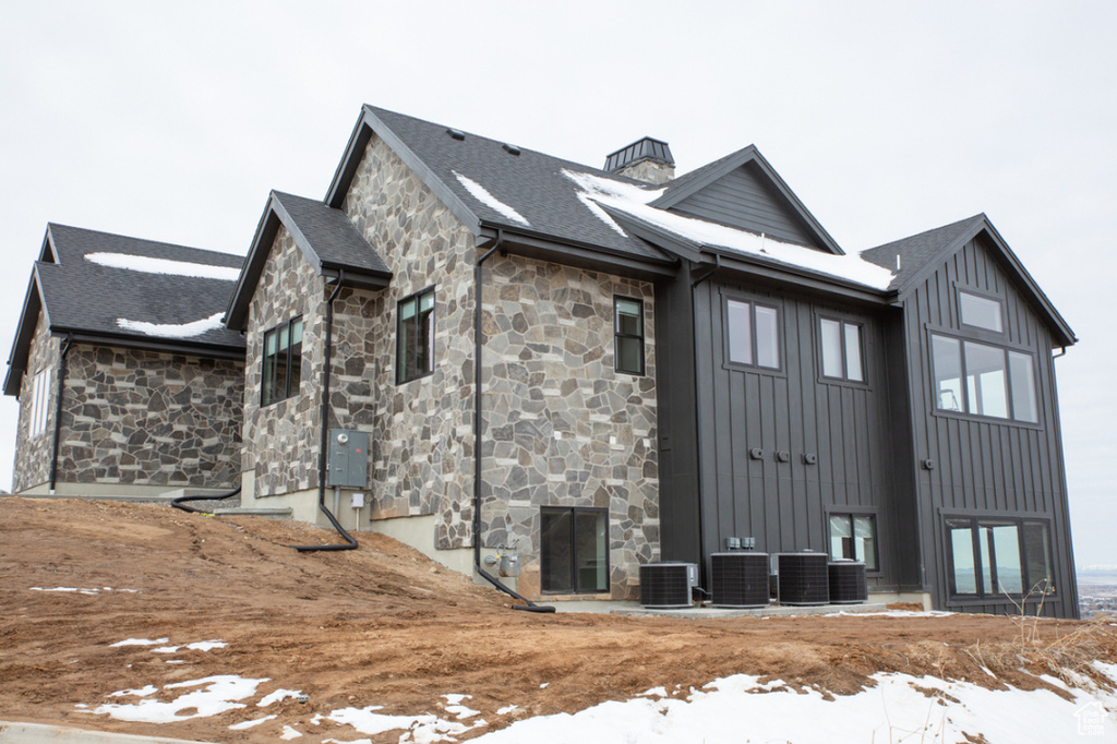 Snow covered property with stone siding, central air condition unit, board and batten siding, and a chimney
