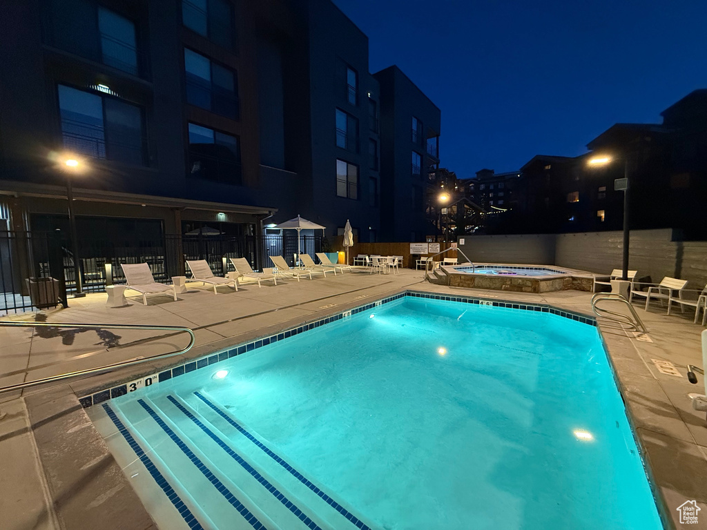 Pool at night featuring a hot tub, a community pool, a patio area, and fence