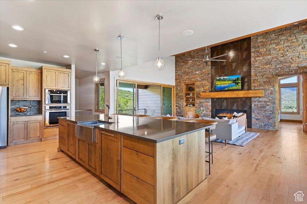 Kitchen with dark countertops, light wood-type flooring, a stone fireplace, an island with sink, and a sink