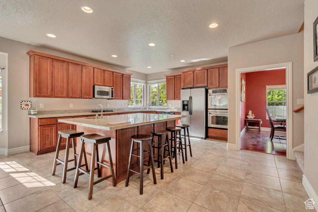 Kitchen featuring appliances with stainless steel finishes, recessed lighting, a center island with sink, brown cabinetry, and plenty of natural light
