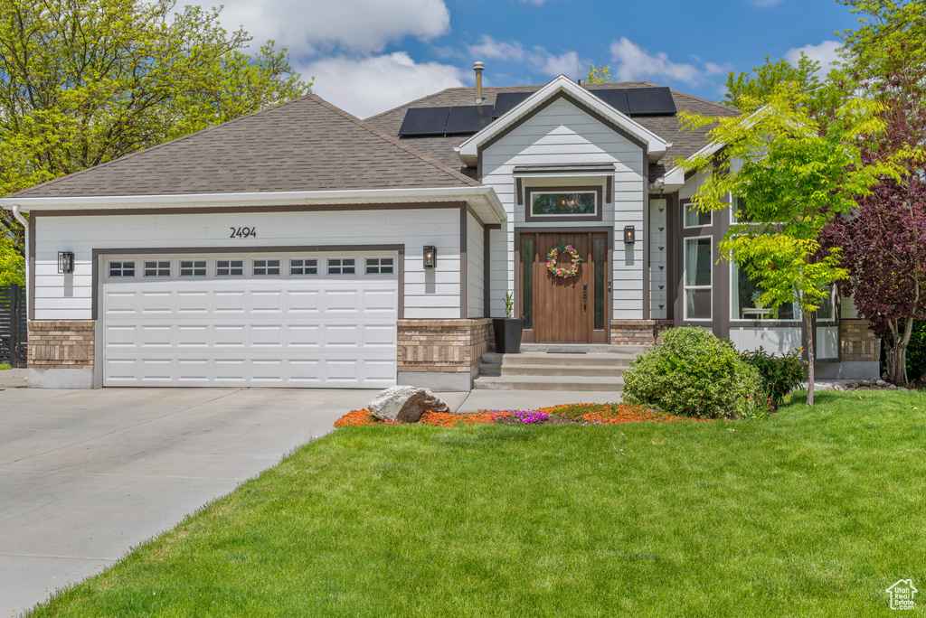 View of front of house with driveway, a front yard, an attached garage, a shingled roof, and solar panels
