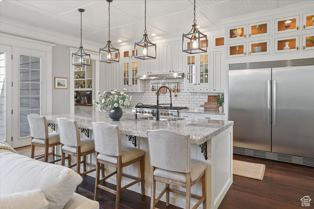 Kitchen with built in refrigerator, tasteful backsplash, white cabinets, dark wood-type flooring, and a center island with sink