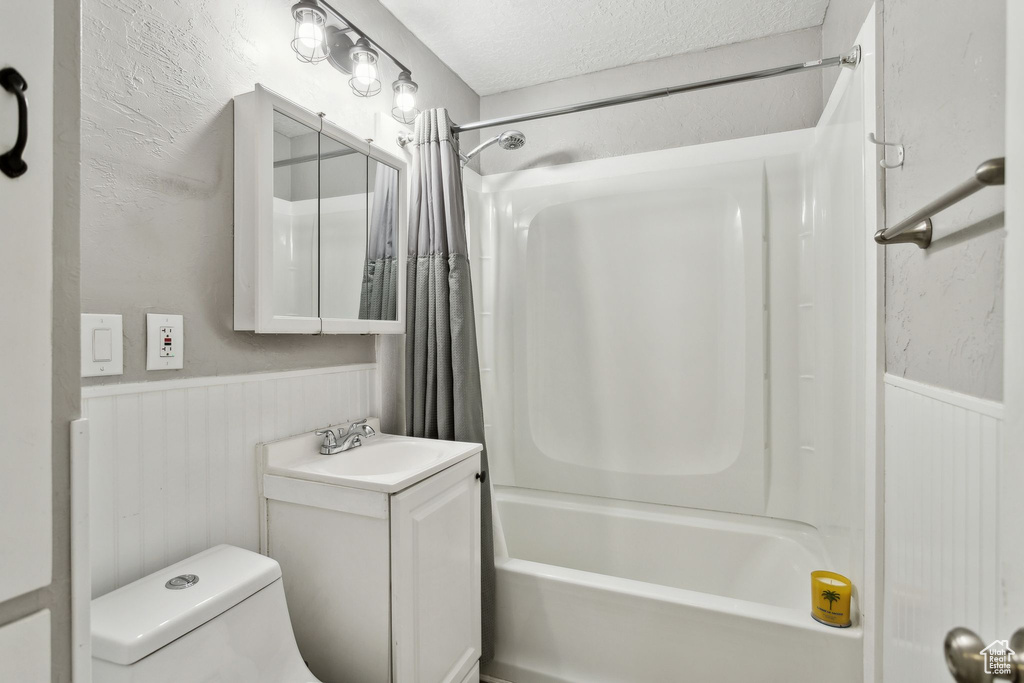 Bathroom featuring a wainscoted wall, shower / tub combo with curtain, toilet, vanity, and a textured ceiling
