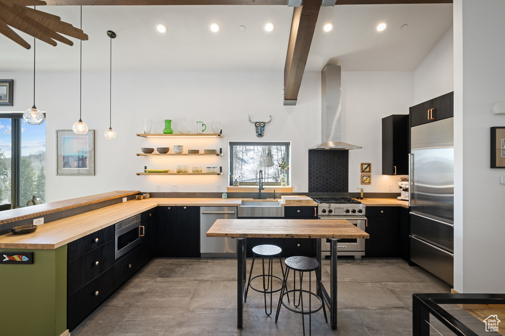 Kitchen with built in appliances, butcher block counters, wall chimney exhaust hood, dark cabinets, and beamed ceiling