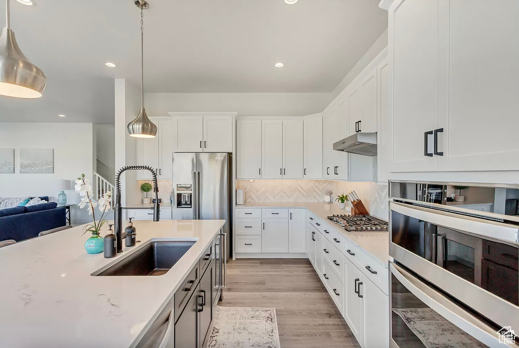 Kitchen featuring a sink, under cabinet range hood, hanging light fixtures, light wood-style floors, and tasteful backsplash