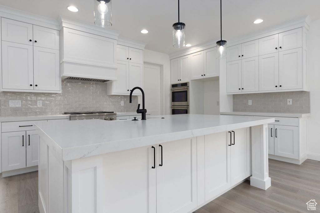 Kitchen with stainless steel double oven, tasteful backsplash, custom exhaust hood, light wood-style flooring, and white cabinetry