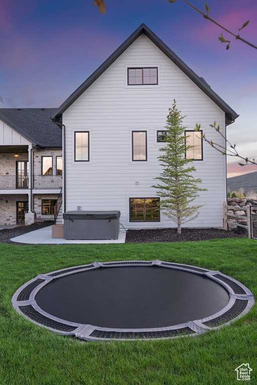 Back of property at dusk featuring a hot tub, a yard, a trampoline, and a patio area