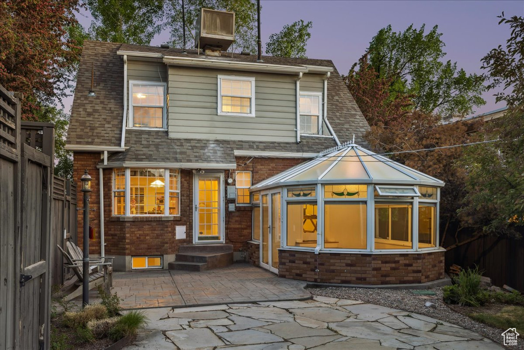 Back of property at dusk with a fenced backyard, brick siding, a patio, a shingled roof, and a sunroom
