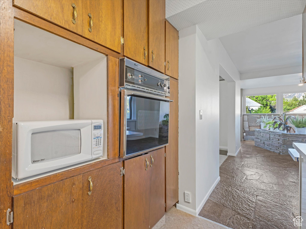 Kitchen featuring white microwave, wall oven, baseboards, brown cabinets, and stone finish flooring