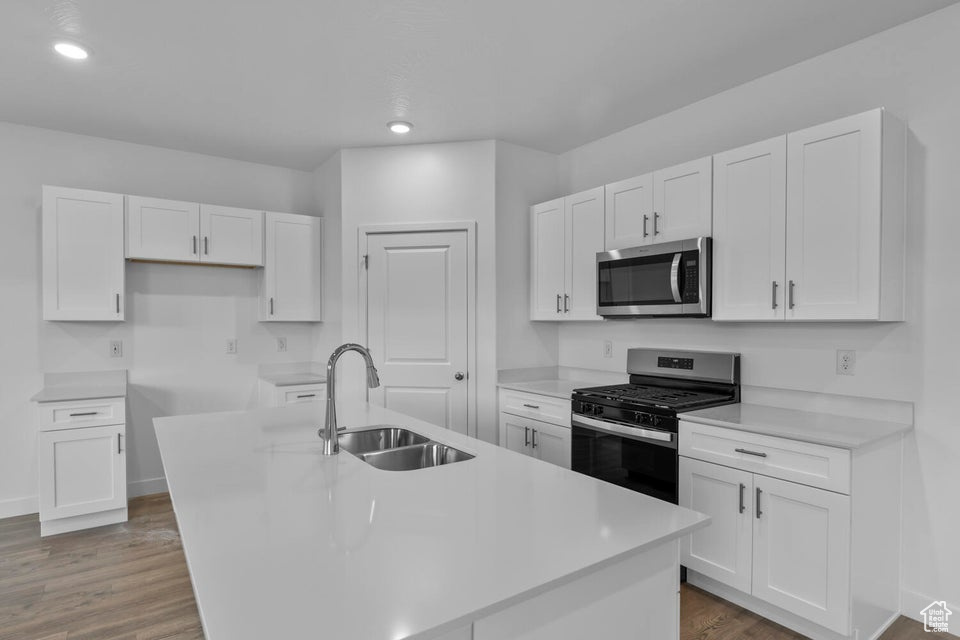 Kitchen featuring appliances with stainless steel finishes, a sink, white cabinets, a center island with sink, and wood finished floors