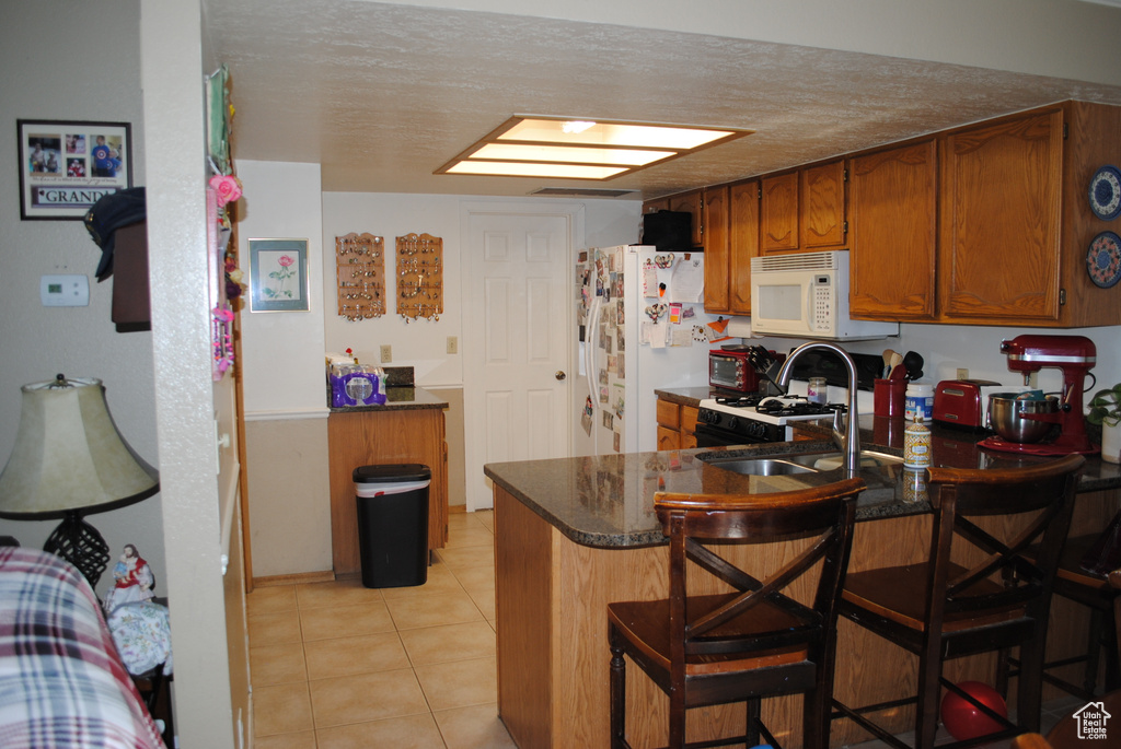 Kitchen featuring white appliances, a peninsula, brown cabinetry, a sink, and light tile patterned flooring