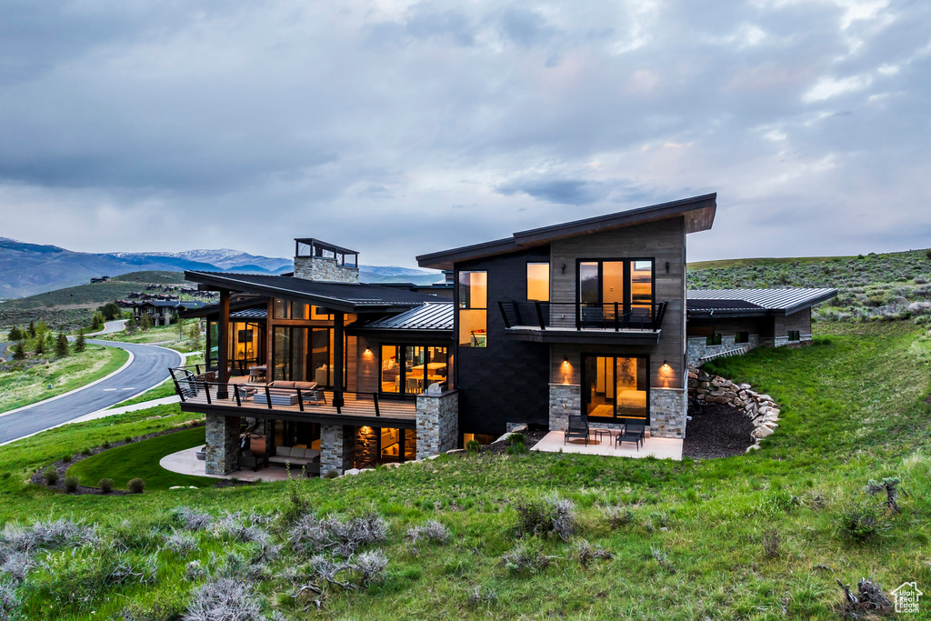 Back of house with a balcony, a patio, a standing seam roof, stone siding, and a yard