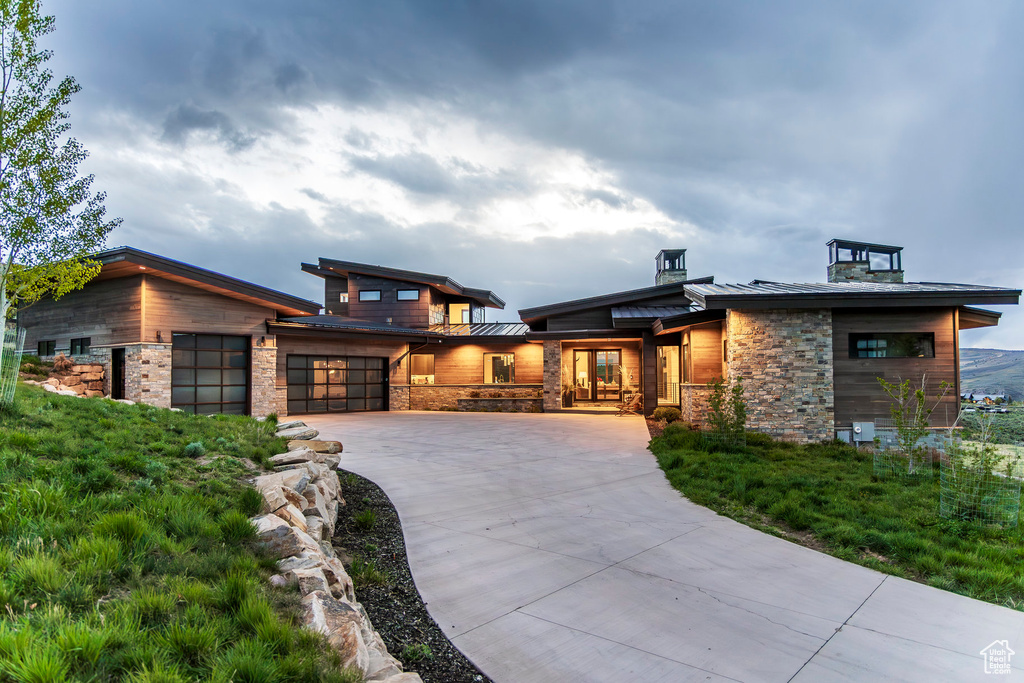 View of front facade featuring a standing seam roof, an attached garage, driveway, metal roof, and stone siding