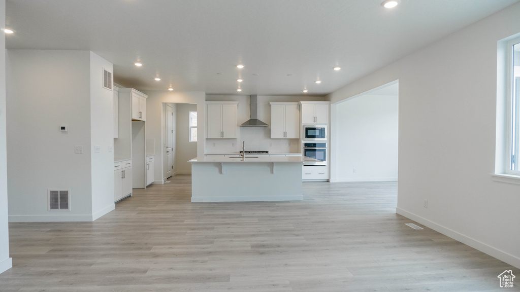 Kitchen featuring wall chimney range hood, open floor plan, white cabinetry, light wood-style floors, and recessed lighting